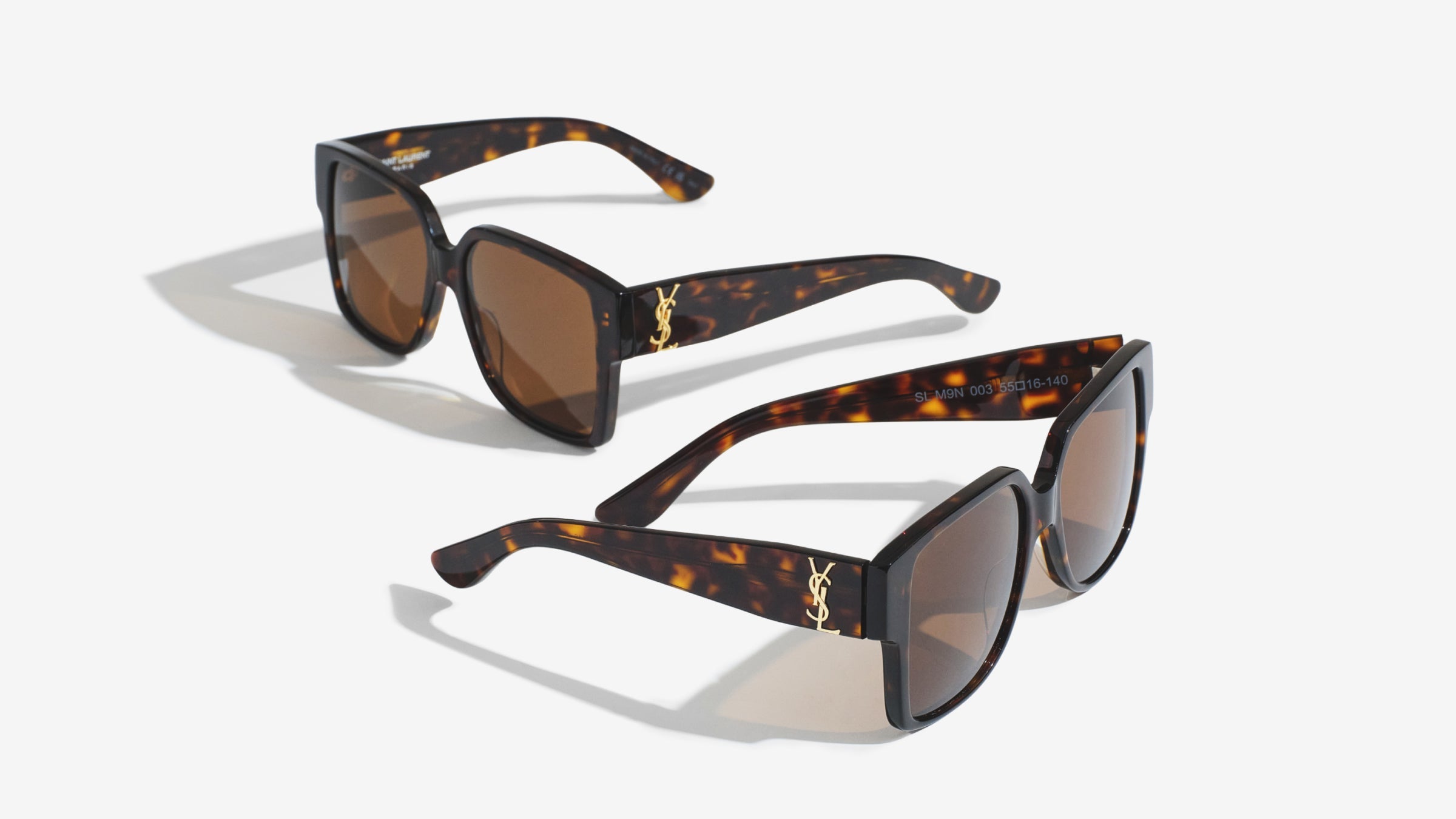 An image featuring two pairs of identical tortoise shell sunglasses with dark brown lenses, arranged on a plain white surface. One pair is positioned slightly behind and above the other, showcasing the gold logo on the side of the thick frames.