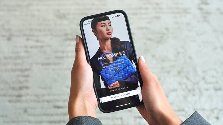 A pair of hands holds a cell phone showing the FASHIONPHILE app sign-up screen