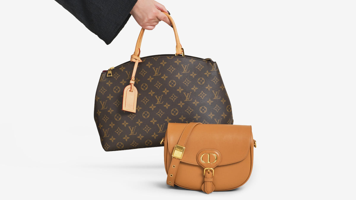 A hand holds an LV Grand Palais bag with a Dior Bobby bag on the floor
