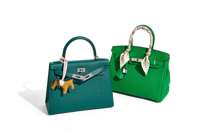 Two green Birkins tilted at an angle, one with a horse charm and one with a scarf wrapped around the handle.