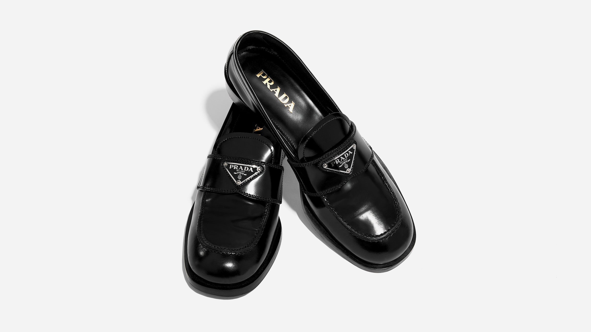 an image of a pair of black spazzolato prada logo loafers styled on a white background by FASHIONPHILE