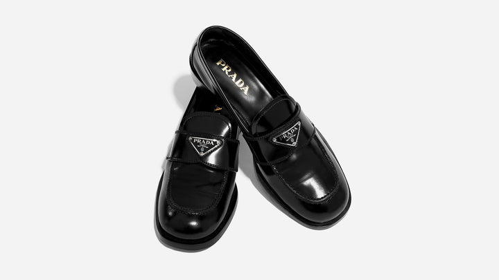 an image of a pair of black spazzolato prada logo loafers styled on a white background by FASHIONPHILE