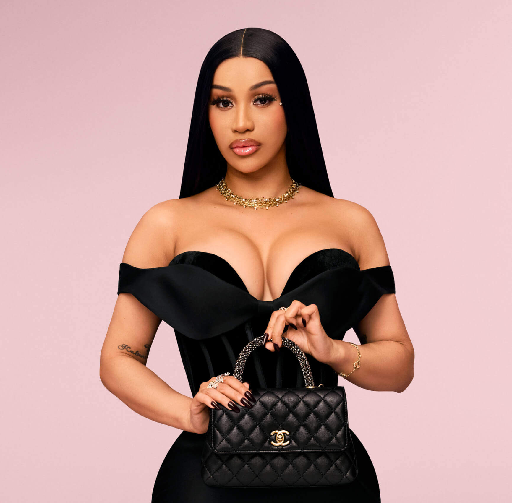 Cardi B with long black hair models a black off-the-shoulder velvet dress and a black quilted Chanel handbag with gold hardware against a solid pink background.